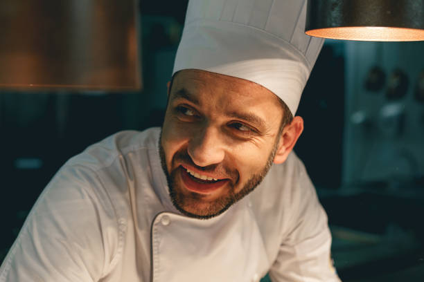 Close up of smiling male chef in uniform standing on kitchen of restaurant and looking away stock photo