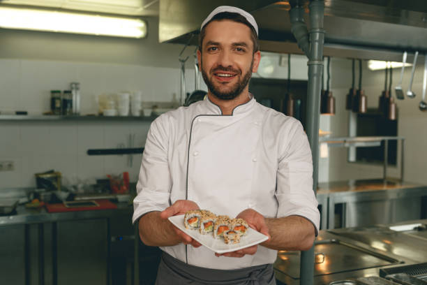 Handsome chef of japanies restaurant showing plate with sushi standing on kitchen stock photo