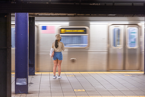 New York Subway, Long Island City, Queens, New York, NY, USA - July 6th 2022: Young woman in summer dress standing in front of a moving subway train at Queens Plaza station