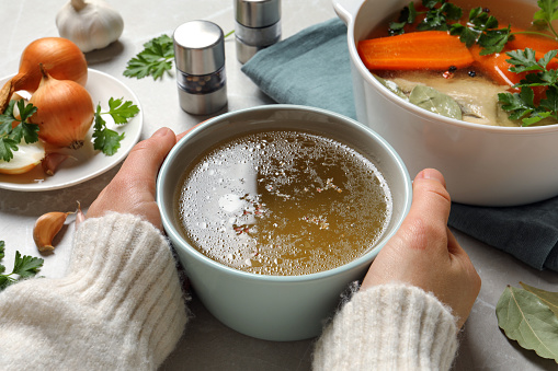 Woman with cup of hot delicious bouillon at light grey table, closeup