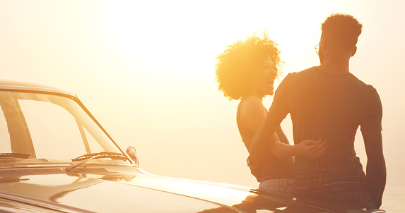 Laughing african couple on sunset road trip bonding, enjoying time together and sharing funny jokes. Smiling, happy or hugging man and afro woman leaning on car on free adventure, holiday or vacation