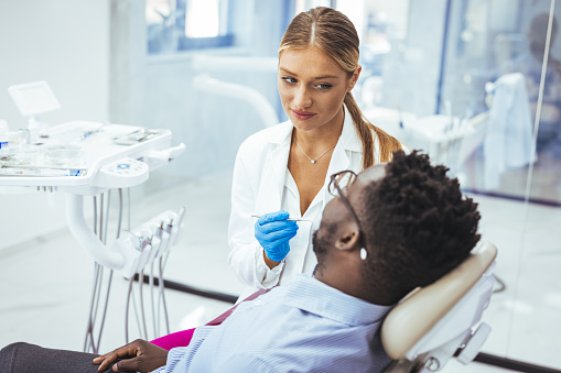 Female dentist with happy male patient at clinic. Happy patient sitting on chair while looking at dentist in medical clinic. Cheerful african american man with during examination in dental clinic