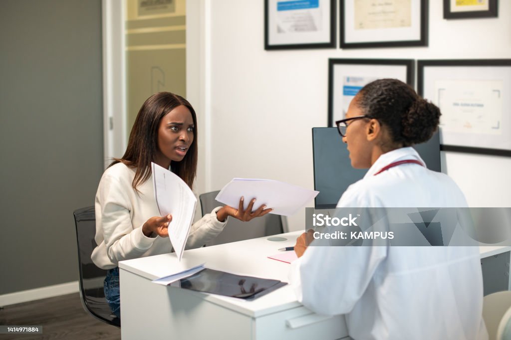 Angry young female patient arguing with doctor about contract Angry young female patient arguing with doctor about contract. African American woman sitting at doctors office exasperated with documents. Medical services contract, medical insurance concept Doctor Stock Photo