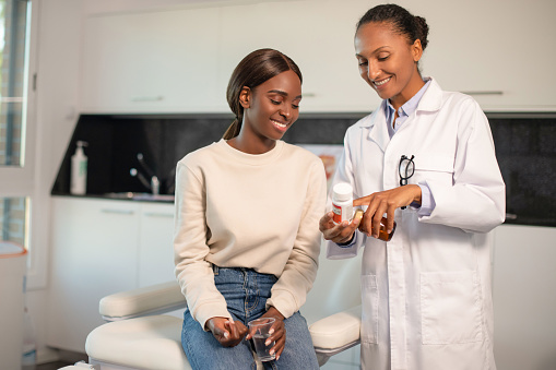 Happy doctor showing vitamins to smiling female patient. Young African American woman sitting at doctors office holding cup of water. Prescription medicine concept