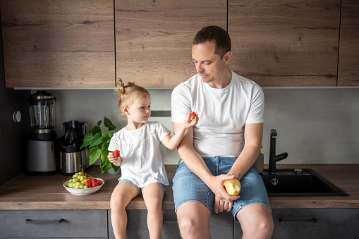 Cute little girl and her handsome dad are eating fruit in modern kitchen. Healthy eating. High quality photo