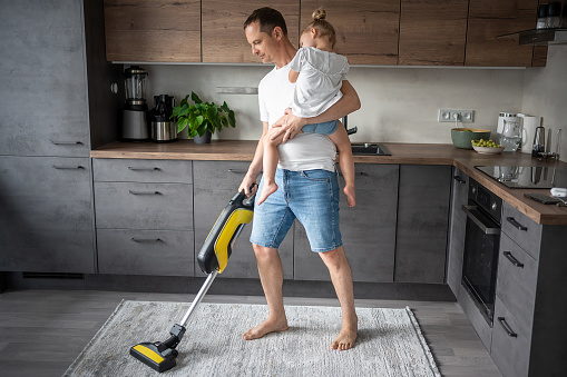 Father with his daughter on his hands vaccuming the floor in the modern home kitchen. High quality photo