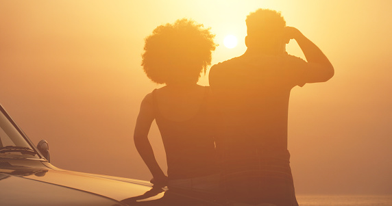 Silhouette of a young couple leaning on a car watching a beautiful, romantic and bright sunset. Loving, relaxing and cute couple enjoying a carefree road trip, getaway or summer vacation on a hot day