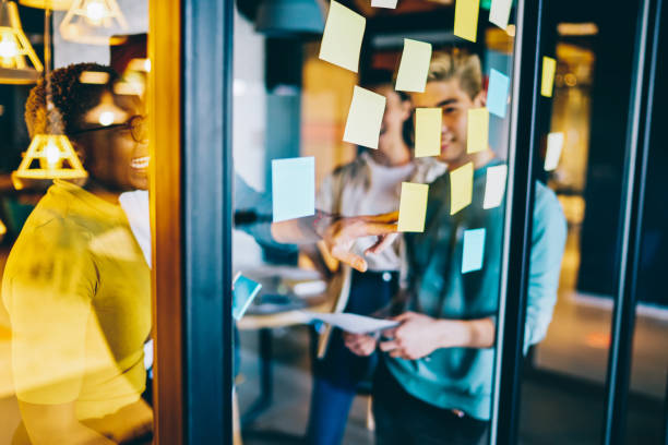 istock Group of happy hipster guys making notes on colorful memo sticks at glass wall while creating interesting ideas, cheerful multiracial male and female smiling while working with startup project 1414974399