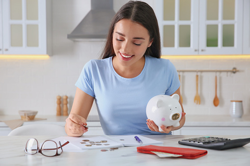 Young woman with piggy bank counting money at table in kitchen