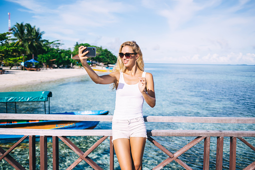 Happy Caucasian tourist in sunglasses using cellphone front camera for making selfie media content with paradise coastline on background, cheerful woman shooting influence video and smiling