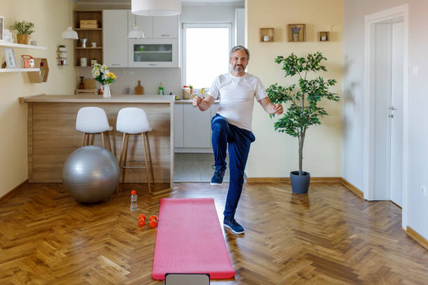 I feel amazing when I excercise A mature Caucasian man is cheerfully looking at the camera while standing with one knee raised up, doing his excercise at home. standing had to knee pose stock pictures, royalty-free photos & images