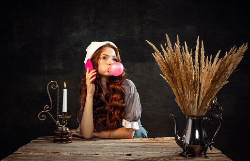 Modern life. Young beautiful redhair girl with long curly hair like girl of renaissance eras isolated on dark background. Comparison of eras, beauty, history, art, creativity.