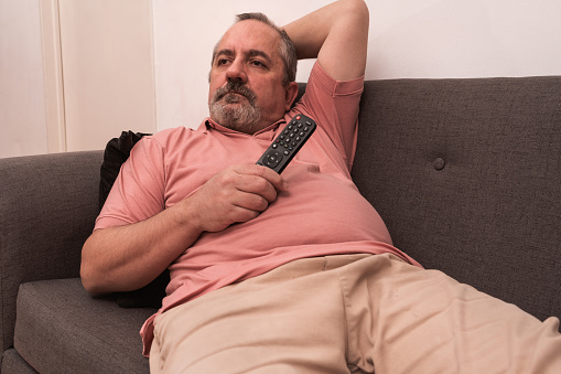 Bearded man lying comfortably on a sofa watching television with the remote control in his hand. Concept of rest, relaxation, home life.