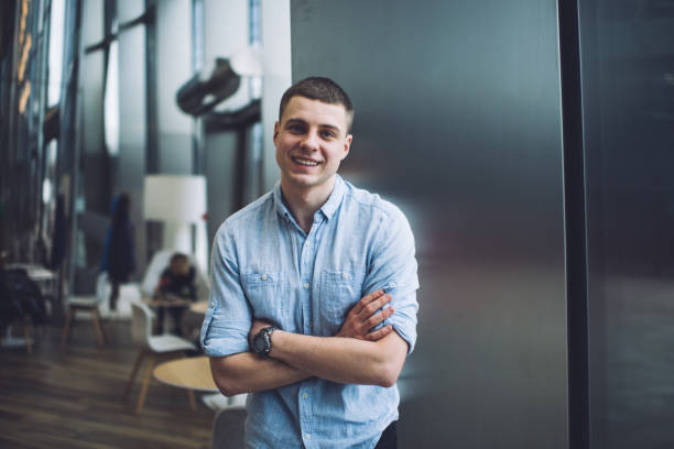 Smiling young male in blue shirt standing with arms folded in spacious office building lobby and looking at camera contentedly