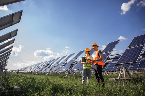 Portrait of engineers spending time outside near solar panels. Two workers: woman and man in special uniform pointing at something and smiling.