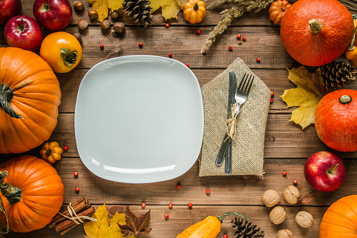 White plate and cutlery surrounded by pumpkins, autumn leaves, apples, kaki persimmon, nuts, cinnamon sticks and cones. Fall flat lay top view composition for Thanksgiving dinner.