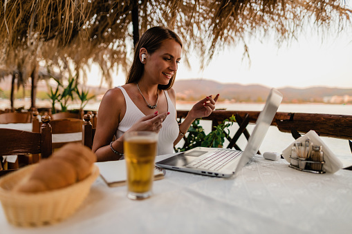 Young woman sitting at the restaurant, using a laptop and headphones having an online class during summer vacation at the seaside