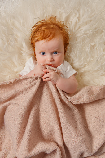 Red hair baby girl is lying with her cuddly toy at home under the blanket. Kid is looking at camera next to rabbit. Cheerful toddler with her favorite toy