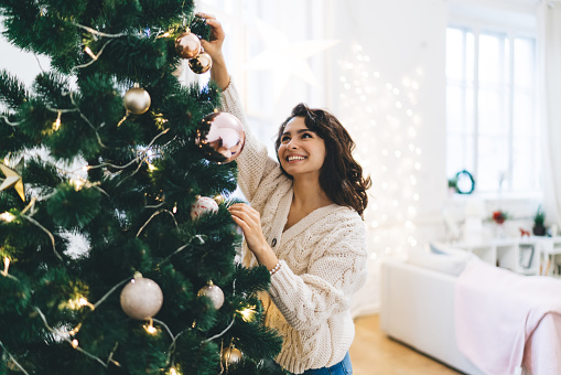 Delight young lady standing near decorated with baubles and garlands Christmas tree on cozy room with star and lights on wall and window