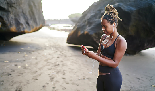 Side view of happy African American female athlete in activewear standing on seashore and surfing smartphone during outdoor training exercises