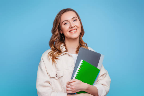 istock Happy girl student in bright clothes holding a notebook, notepad, a folder in her hands smiling and looking at the camera on a pink background, copy space for text 1415017563