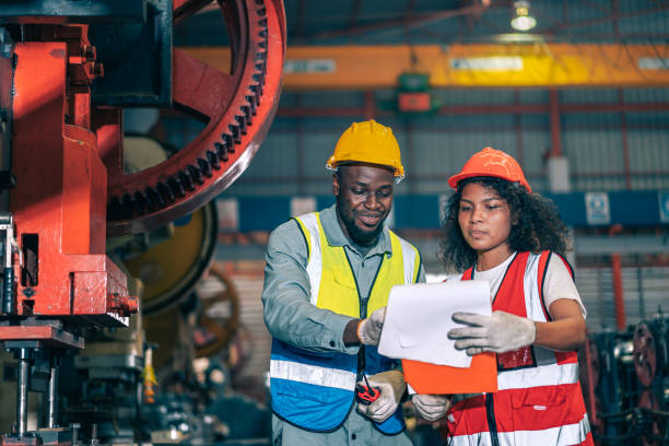 istock Two professional engineer/worker/technician use clipboard discuss work, walk in steel metal manufacture factory plant industry. Black African American man and woman wear hard hat check quality machine 1415018741