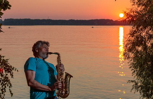 An adult man plays the saxophone at sunset by the river in the evening.