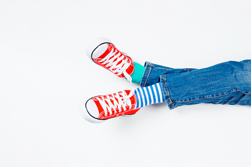 Kid wearing different pair of socks. Child foots in mismatched socks and red sneakers sitting on white background. Odd Socks day, Anti-Bullying Week, Down syndrome awareness concept.
