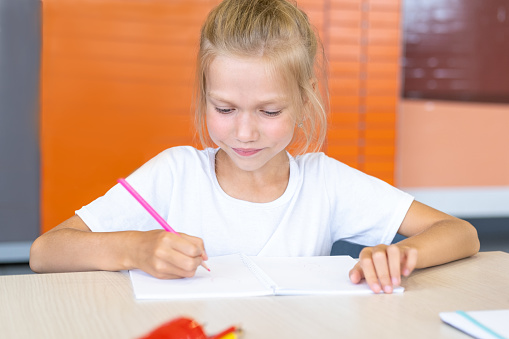 Focused girl doing homework, writing text in a notebook at the table. Cute positive girl writes and smiles. Back to school concept. School and preschool education.