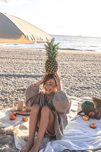 A little girl on a sandy seashore on a blanket at a picnic with fruit.