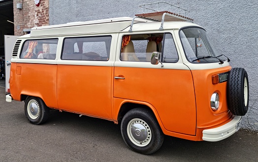 Front and side view of an Orange and White Volkswagen T2 1600cc camper van built in Brazil. This VW campervan is parked on tarmac by a wall.