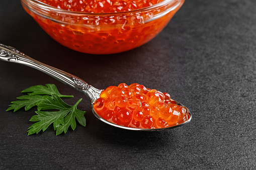 Salmon red caviar in a spoon on a black slate background. Vintage spoon full of trout caviar and parsley leaf close-up. Seafood fish delicacy. Salted salmon roe for gourmet food concept. Front view.