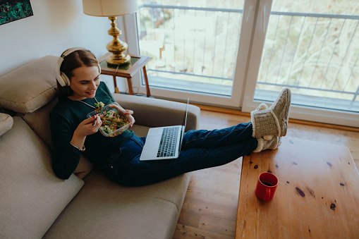 Young woman sitting on the couch in the living room, using wireless headphones and a laptop, eating a healthy salad, and watching streaming services at home