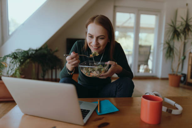 istock Young woman eating a salad and using a laptop at home 1415053210