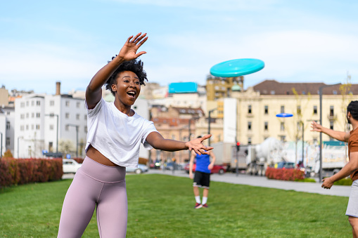 Young Woman of African-American Ethnicity is Playing with Frisbee in Public Park and Trying to Catch it.