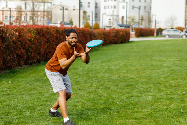 Young African Man is Catching a Frisbee while Playing with Friends in a Park. Young Man of African-American Ethnicity is Playing with Frisbee in Public Park and Trying to Catch it. plastic disc stock pictures, royalty-free photos & images