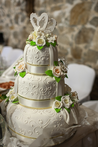 A bride and groom, who are wheelchair users that have just got married, at their evening reception in a wedding venue in Morpeth, North East England. They are out of focus, looking at the camera while smiling behind their wedding cake. The cake is in focus and has figurines of the happy couple made out of icing on top of it.