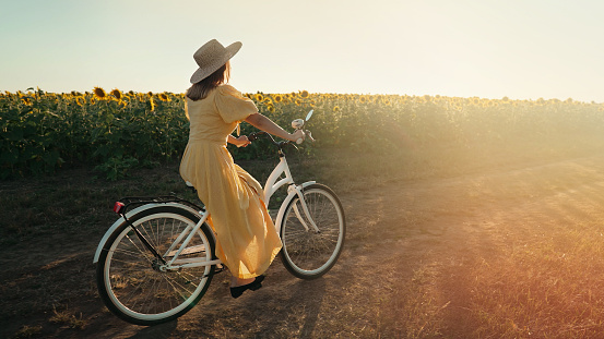 Rural woman in timeless dress riding retro styled white bicycle on country road alone near sunflowers field. Vintage fashion, amazing adventure, countryside activity, healthy lifestyle. quality photo