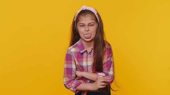 Cheerful funny young preteen child girl kid showing tongue making faces at camera, fooling around, joking, aping with silly face, teasing. Little toddler children isolated on studio yellow background