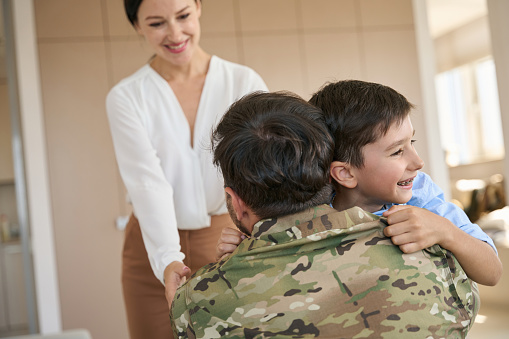 Military father in camouflage clothes tightly hugging his teenager son and beautiful elegant wife in a bright room