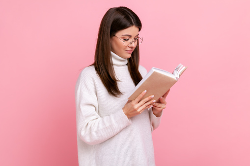 Concentrated smiling brunette girl holds book, enjoys reading, standing absorbed with exciting plot, wearing white casual style sweater. Indoor studio shot isolated on pink background.