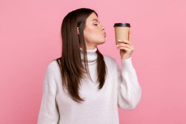 Side view of female with dark hair kissing disposable cup of take away coffee, enjoying beverage. Side view of female with dark hair kissing disposable cup of take away coffee, enjoying beverage, wearing white casual style sweater. Indoor studio shot isolated on pink background. coffee addict stock pictures, royalty-free photos & images