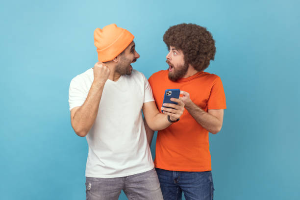 dos hombres asombrados emocionados de pie con el teléfono celular y celebrando el éxito, apretaron los puños. - super powers fotografías e imágenes de stock