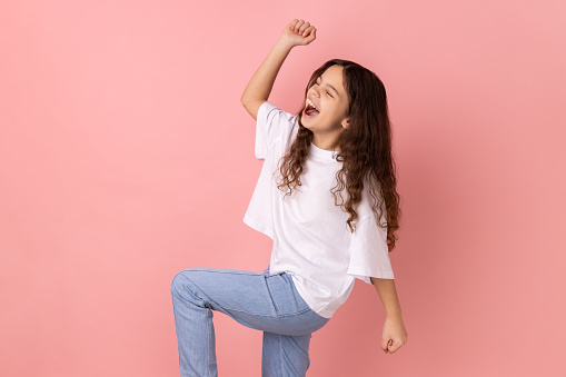 Happy satisfied little girl wearing white T-shirt showing yes gesture and screaming celebrating her victory, success, dreams comes true, euphoria. Indoor studio shot isolated on pink background.