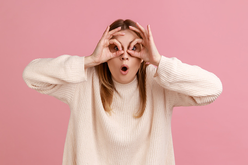 Portrait of astonished blond woman spying, watching unbelievable shocking event, looking through fingers imitating binoculars, wearing white sweater. Indoor studio shot isolated on pink background.