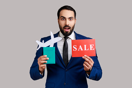 Surprised bearded man holding passport with paper air plane and sale word sign, expressing astonishment, traveling, wearing official style suit. Indoor studio shot isolated on gray background.