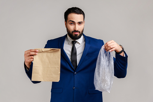 Portrait of confused puzzled bearded man holding paper and plastic package in hands, decides what to use, wearing official style suit. Indoor studio shot isolated on gray background.