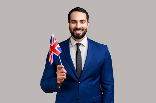 Delighted bearded man holding British flag, celebrating holiday, looking at camera with toothy smile, wearing official style suit. Indoor studio shot isolated on gray background.