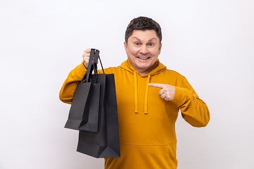 Smiling middle aged man pointing finger at black paper bags with purchases, satisfied with shopping, seasonal sale, wearing urban style hoodie. Indoor studio shot isolated on white background.