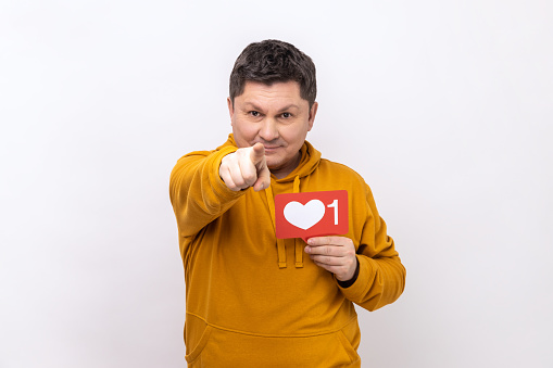 Portrait of happy positive man showing blogger heart icon and pointing to camera, asking to click button, wearing urban style hoodie. Indoor studio shot isolated on white background.